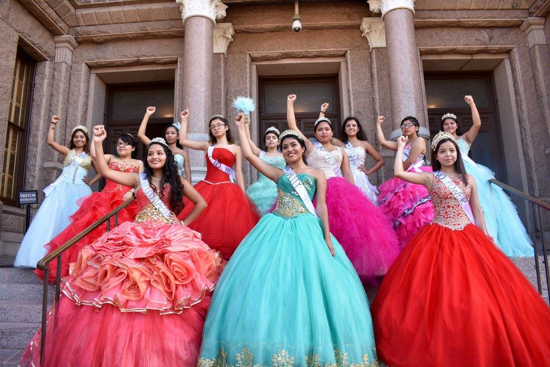 12 young Latina women in quinceañera dresses stand on the stairs of a government building with their fists raised.