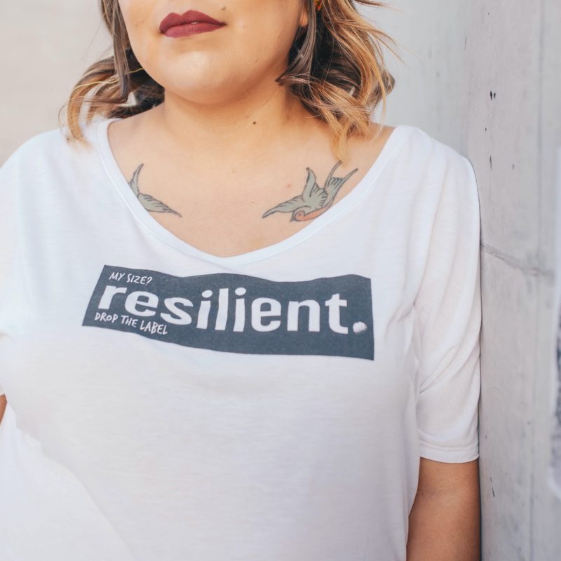 Woman with light hair and tattoos leaning on white wall wearing a shirt that says "resilient".