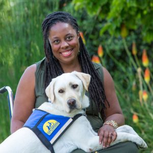 India Harville, African American female with long black locs, seated in her manual wheelchair wearing a long sleeveless green dress. Her service dog, Nico, a blond Labrador Retriever, has his front paws on her lap. He is wearing a blue and yellow service dog vest. They are outside with greenery behind them.