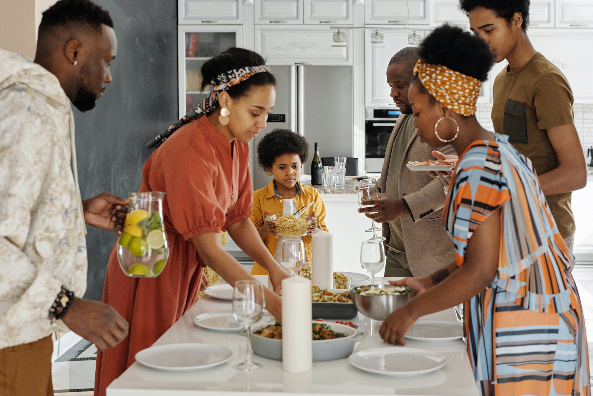 A Black family made up of two men, two women, a young adult, and a child set the table for a meal.