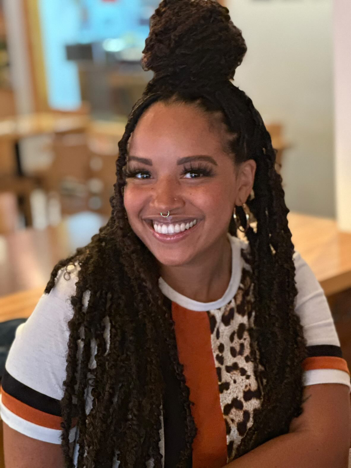 Kiara Lee-Benton is smiling with hair half up in a bun and the rest cascading down her shoulders. She is wearing a white shirt with black and brown stripes and leopard print.