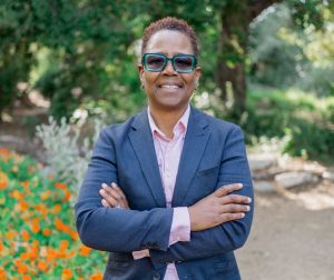 Darlene Nipper stands in a garden in arms casually crossed, smiling, and wearing blue sunglasses, a blue suit, and pink collared shirt.